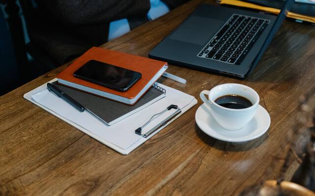 Laptop, clipboard, notebook and a coffee cup on a wooden desk