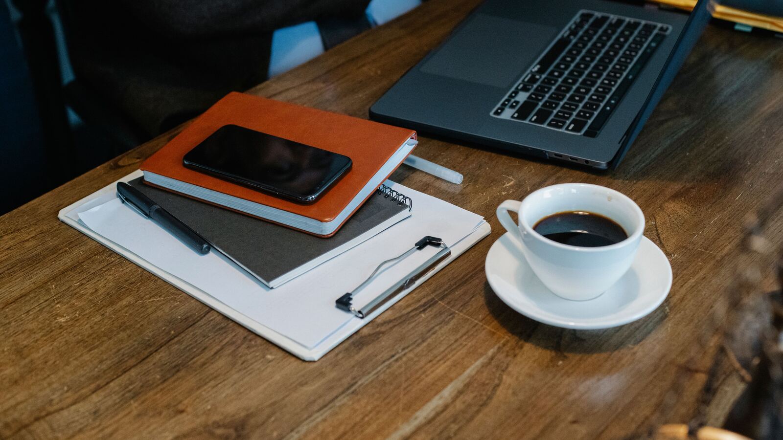 Laptop, clipboard, notebook and a coffee cup on a wooden desk